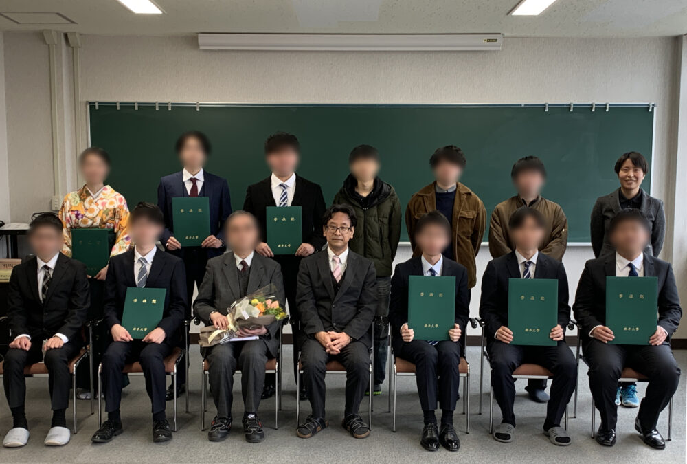 前列のかたは椅子に掛け、後列のかたは立ち、卒業証書を手にしたり、花を持ったりして、卒業時の記念撮影